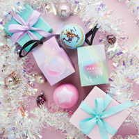 Miss Patisserie joy bath ball. Image shows the box, the unboxed bath ball, some wrapped presents, the Peace bath ball and lots of Christmas decorations such as tinsel. Vegan and cruelty-free. Available at Lovethical along with plenty of other vegan and cruelty-free beauty products, makeup, make up, toiletries and cosmetics for all your gift and present needs. 