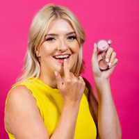 Pura Cosmetics pink tinted lip balm. Vegan and cruelty-free. Image shows a smiling person holding the lip balm and applying it to their lips. Available at Lovethical along with plenty of other vegan and cruelty-free beauty products, makeup, make up, toiletries and cosmetics for all your gift and present needs. 