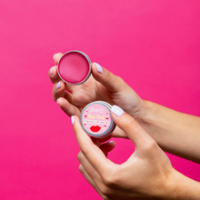 Load image into Gallery viewer, Pura Cosmetics pink tinted lip balm. Vegan and cruelty-free. Image shows a person holding the tin open and showing the vibrant pink colour within. Available at Lovethical along with plenty of other vegan and cruelty-free beauty products, makeup, make up, toiletries and cosmetics for all your gift and present needs. 
